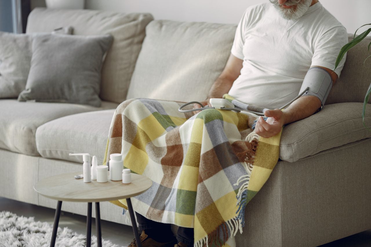 Services Elderly man monitoring blood pressure on a cozy couch at home, surrounded by medication.