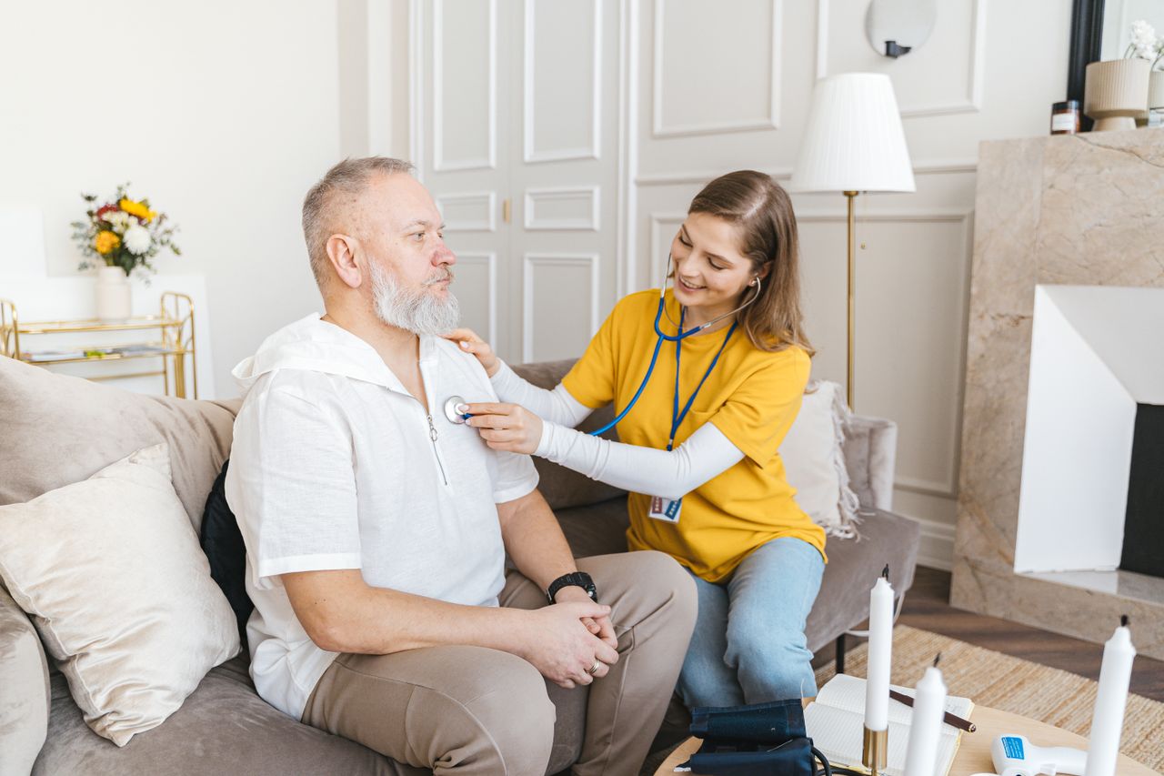 Services A nurse uses a stethoscope for a home check-up on a senior adult in a cozy living room.